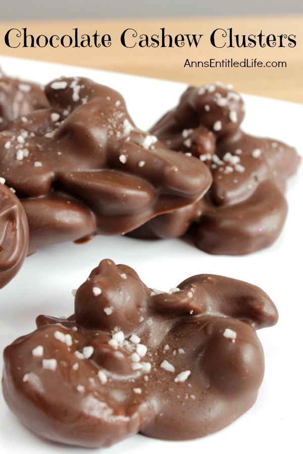 A homemade chocolate cashew cluster is front and center. A small pile of additional chocolates are in the background. These are set on a white paper.