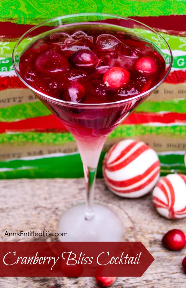 A martini glass filled with a cranberry cocktail standing against a red and green background. There are red and white holiday balls in the lower right.