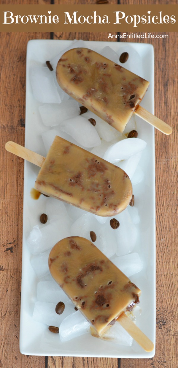 Three homemade brownie mocha popsicles in a vertical line on top of a bed of ice on a white plate, with a few coffee beans resting on the white plate too. This is on top of a brown board.