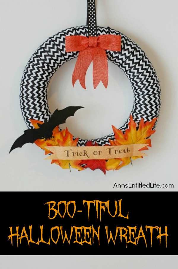 A Halloween wreath done in black and white ribbon, with a bat, bow, and leaves accent, hanging on a white wall.