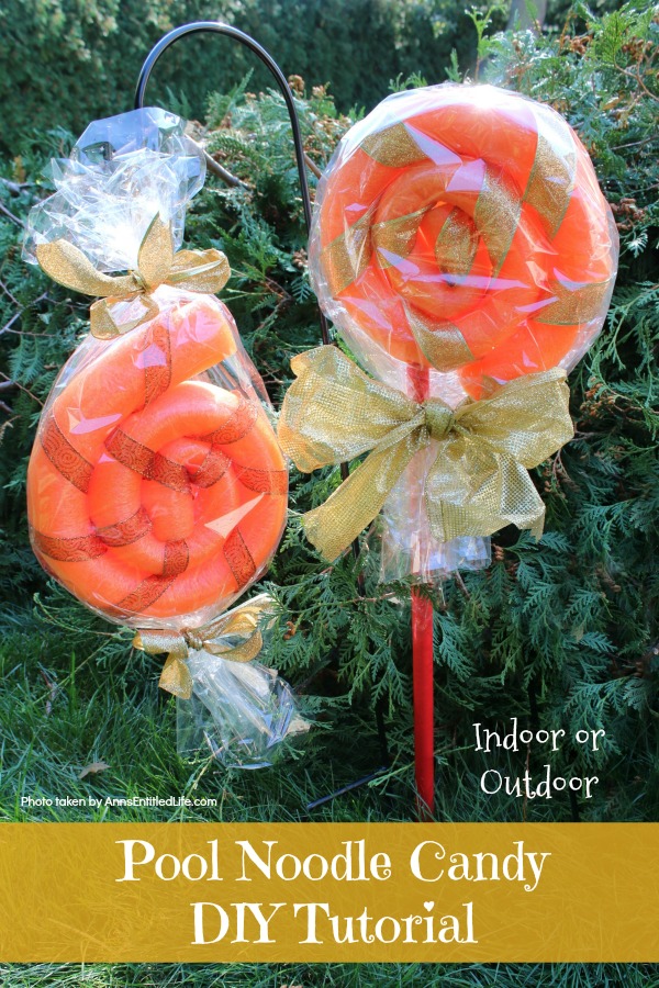 Two orange pool noodles formed to look like a candy, covered in cellowrap, one with red pvc piping against an evergreen background 