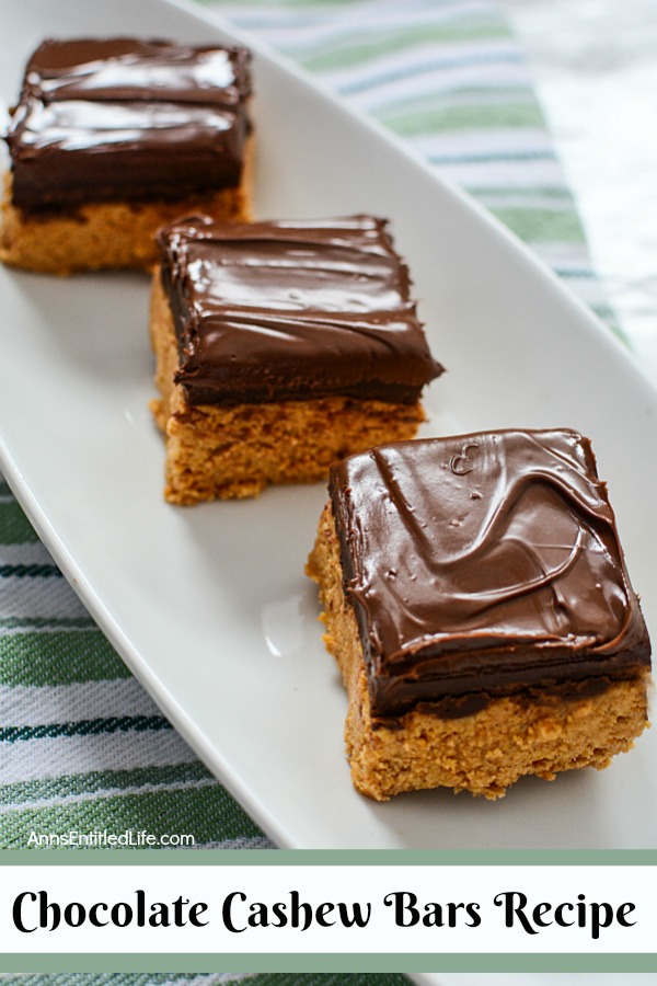 three cashew cookie bars topped with chocolate on a white serving dish on top of a green striped dishtowel