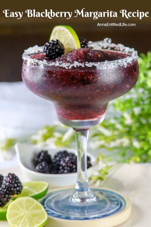 Side view of a blackberry margarita in a glass, a lime and fresh blackberry garnish on a skewer lays across the back. Below is a bowl of fresh blackberries, and more sliced limes