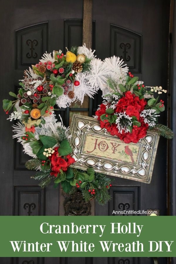 A large white wreath decorated with a sign, holly berries, and faux greenery handing on a bronze wreath hanger against a dark door.