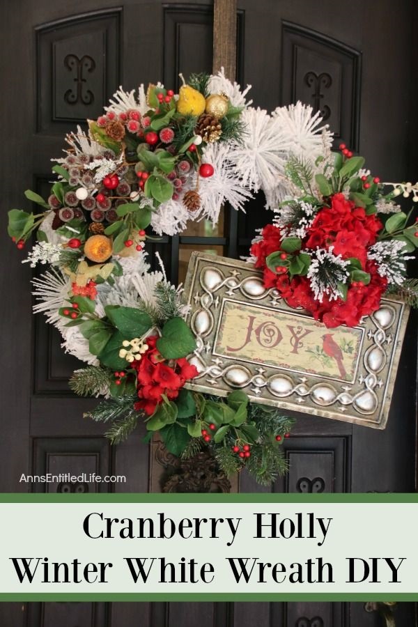 A large white wreath decorated with a sign, holly berries, and faux greenery handing on a bronze wreath hanger against a dark door.
