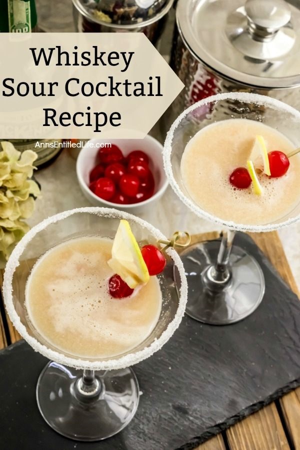 An overhead shot of two cocktail glasses filled with whiskey sour, a white bowl filled with cherries above them, a cocktail shaker in the background