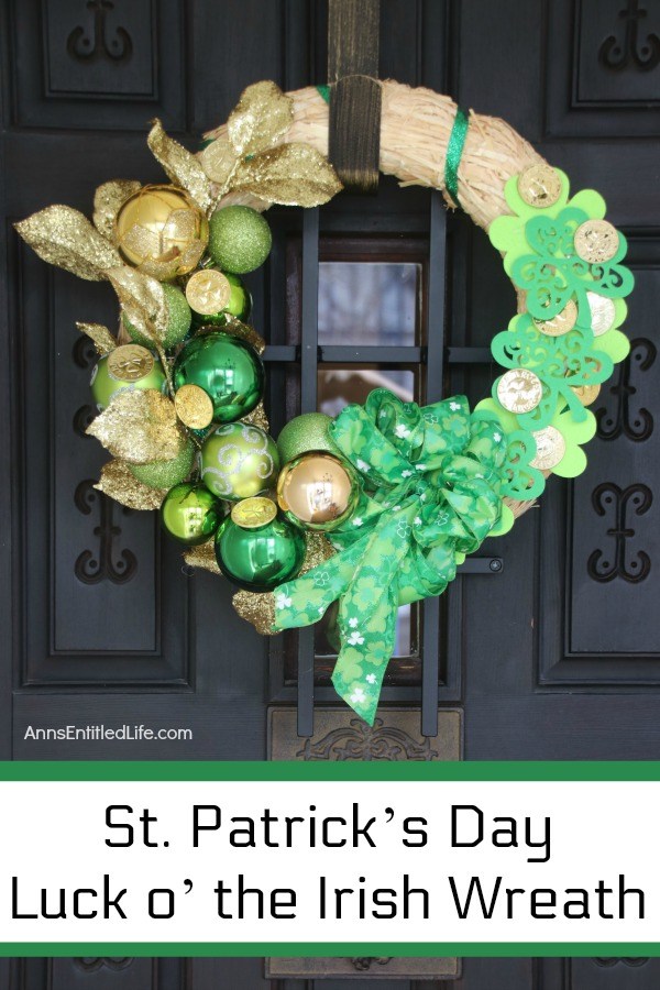 straw wreath decorated for st patrick's day with green and gold bulbs, green ribbon, and gold coins, hanging on a bronze door hanger on a dark colored door