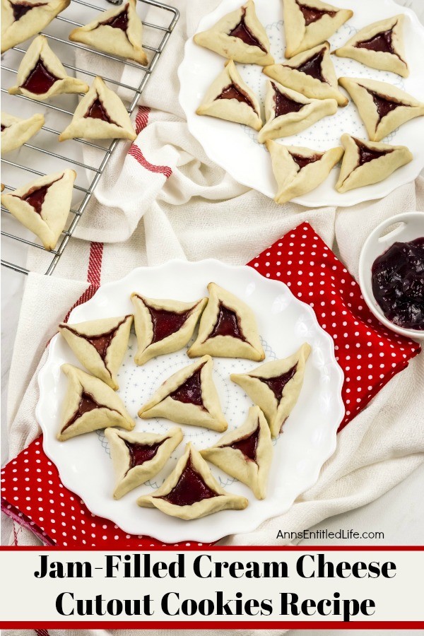 An overhead view of two white plates willed with jam cookies (triangular shaped cookies with jam in the middle). A rack has more jam cookies in the upper left. There is a bowl of jam in the middle right.
