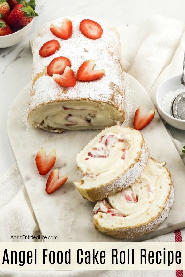  An overhead view of an angel food cake roll on a marble cutting board, two pieces are cut and laying in front of it. There are two bowls on the right filled with strawberries and cream