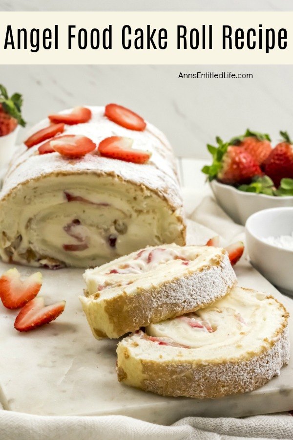 Angel food cake roll on a marble cutting board, two pieces are cut and laying in front of it. There are two bowls on the right filled with strawberries and cream