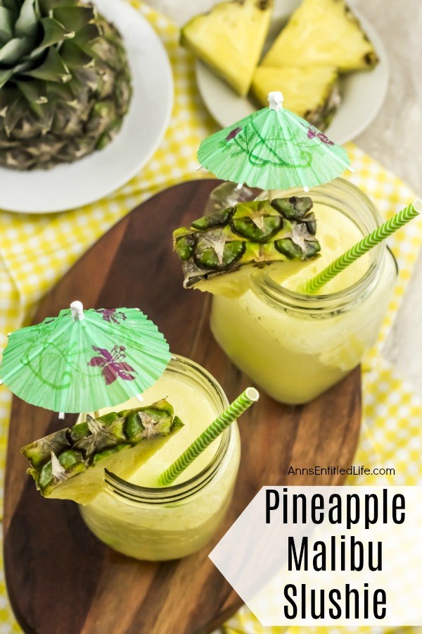 2 yellow pineapple cocktails on a brown wooden board, with fresh pineapple garnish and decorative drink umbrellas. Cut up fresh pineapple in the background