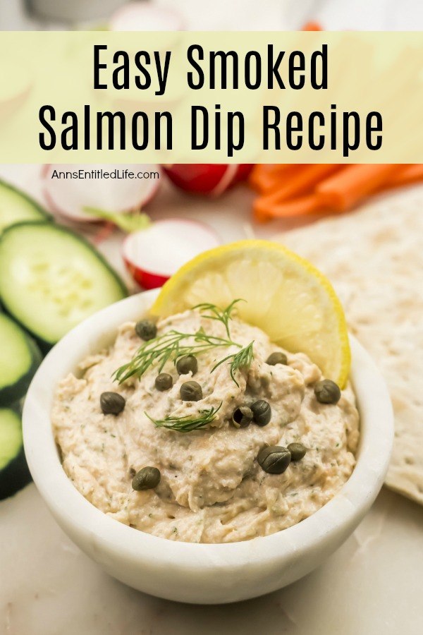 salmon dip in a white bowl garnished with capers, dill and lemon, sliced cucumbers, radishes, and carrots in the background with crackers to the right of the bowl