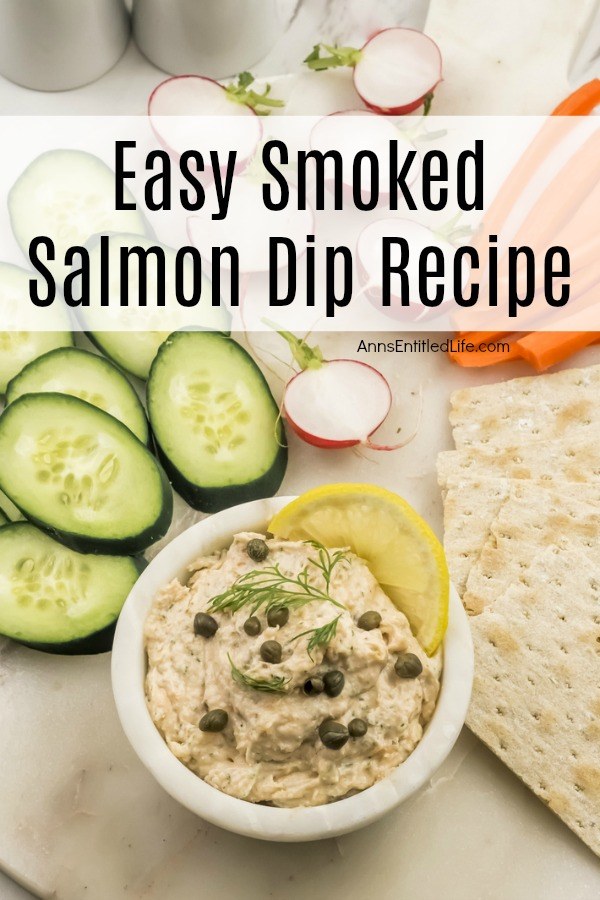 salmon dip in a white bowl garnished with capers, dill and lemon. There are sliced cucumbers, radishes, and carrots in the background along with crackers