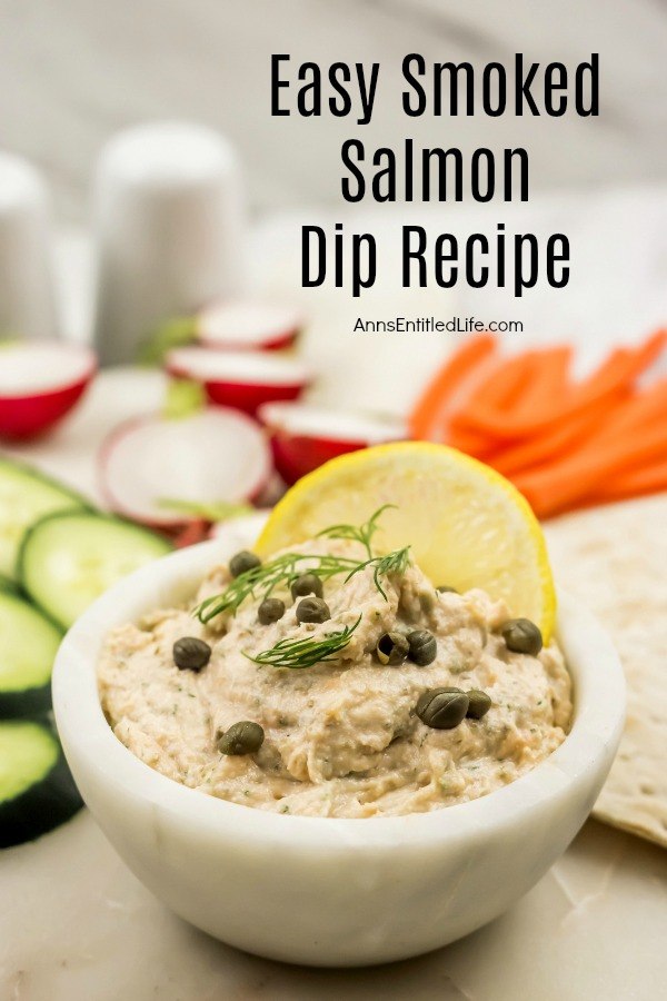 salmon dip in a white bowl garnished with capers, dill and lemon, sliced cucumbers, radishes, and carrots in the background with crackers to the right of the bowl