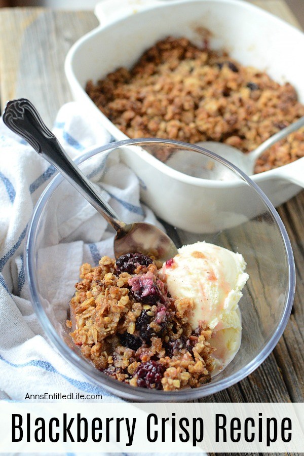 a serving of blackberry crisp a la mode with ice cream in a clear bowl and spoon, blue striped napkin to the left, the serving container and serving spoon of blackberry crisp is in the background