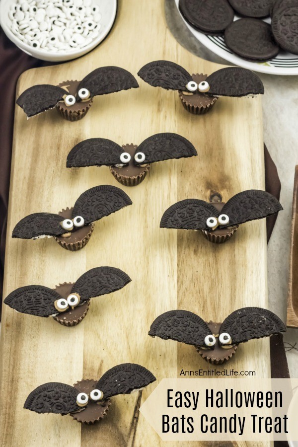7 candy treats made up to look like bats, on a butcher block cutting board, a plate of cookies in the upper right, a bowl of candy eyes in the upper left
