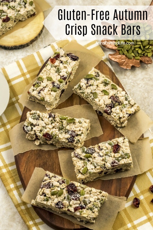 5 Gluten-Free Autumn Crisp Snack Bars served on small pieces of butcher paper, on a brown cutting board atop a yellow and white dish towel, a dish of pepitas is in the upper right, another snack bar sits on a wooden tray in the upper left