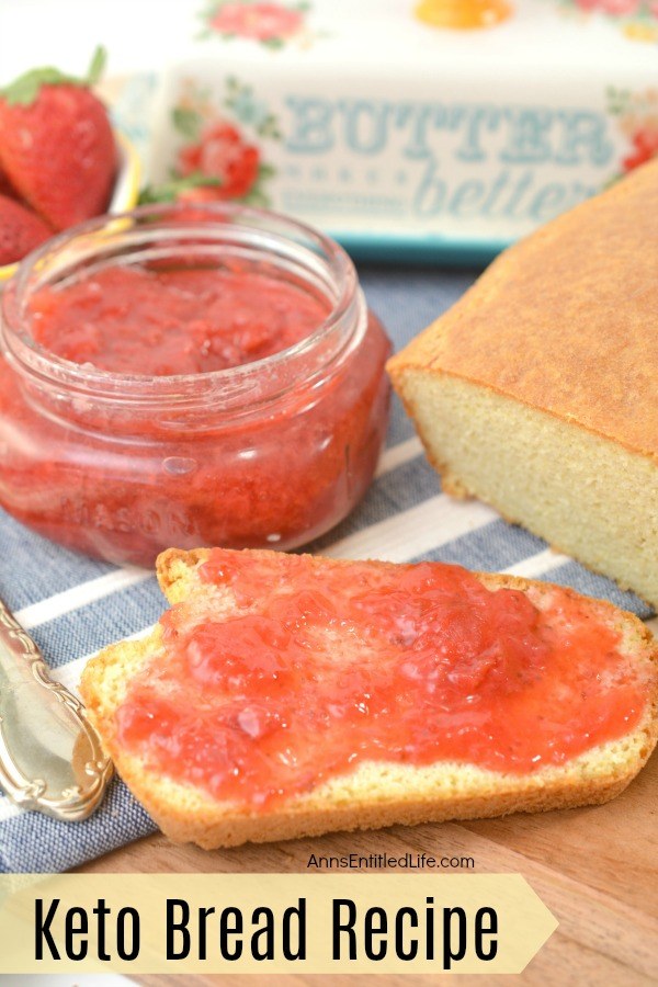 keto bread and keto strawberry jelly half on a butcher block board and half on a grey and white towel, the jar of jelly in slightly to the upper left, the bread load is slightly to the upper right, a butter dish in the center top. A dish of fresh strawberries are to the jelly's upper left, and a serving utensil is to the left of the bread and jelly.