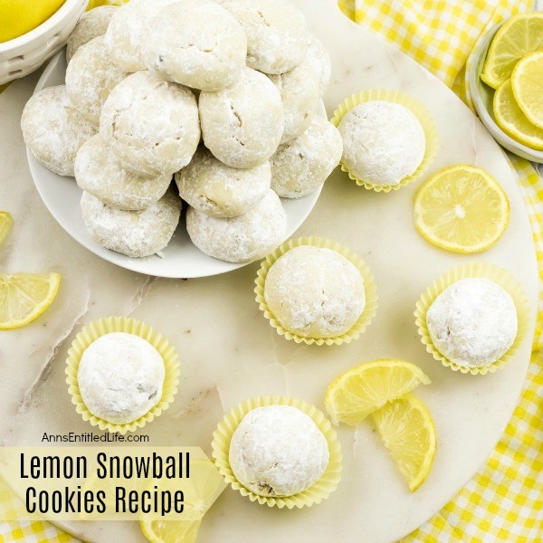 5 lempon snowball cookies in yellow cupcake holders, a white plate full of lemon snowball cookies in the upper left. Fresh lemons are displayed as garnish. All sits on a white board