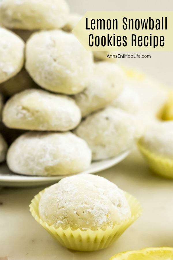 a lemon snowball cookie in yellow cupcake holder is featured in front of a white plate full of lemon snowball cookies in the background. Fresh lemons are displayed as garnish. All sits on a white board