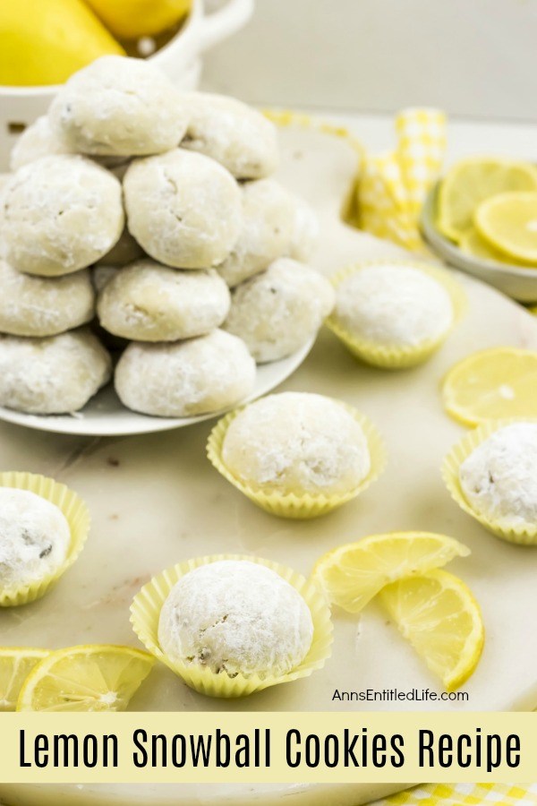 5 lempon snowball cookies in yellow cupcake holders, a white plate full of lemon snowball cookies in the upper left, a basket of lemons behind the plate. Fresh lemons are displayed as garnish. All sits on a white board