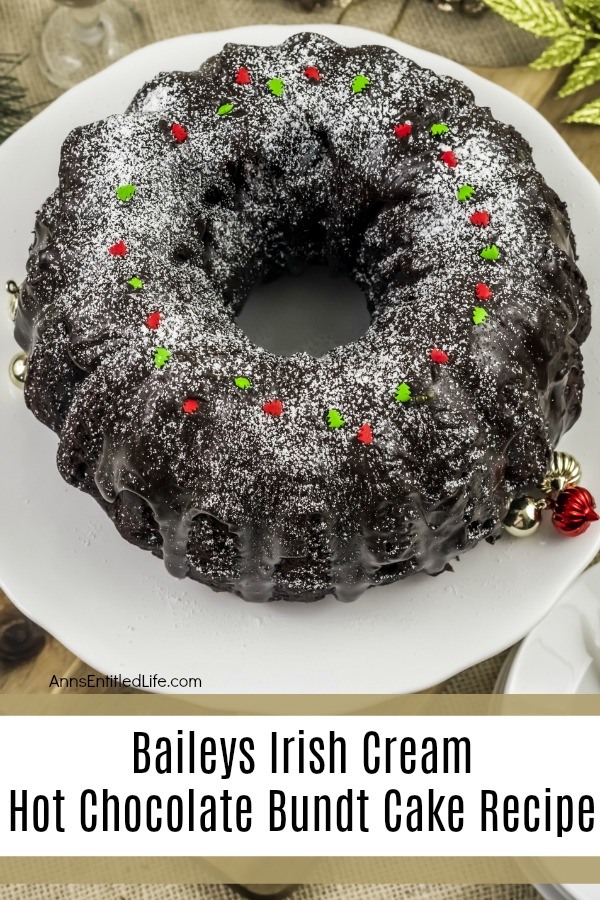 A whole Baileys Irish Cream Hot Chocolate Bundt Cake sitting on a white platter. There is a stack of white dishes in the lower right, some holiday decor in the upper left.