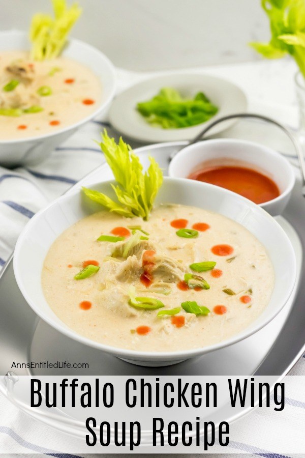 A white bowl filled with Buffalo chicken wing soup set in a silver tray, a small white bowl of hot sauce next to it, a second bowl is in the upper right, a bowl of celery stalks in the upper middle.