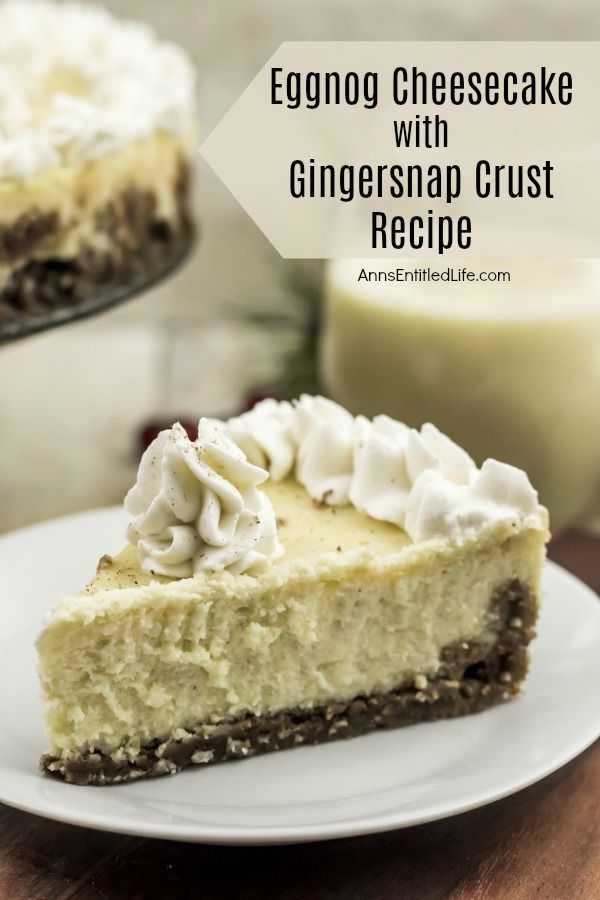 An extreme closeup of a slice of eggnog cheesecake on a white plate is front and center, the remaining eggnog cheesecake sits in the upper left. There is a glass of eggnog in the upper right.