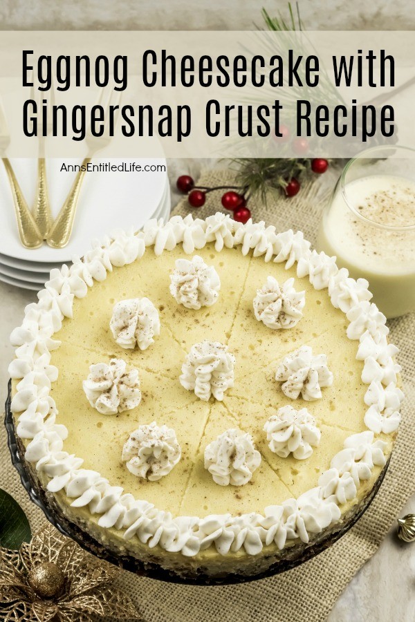 A fully decorated eggnog cheesecake sits in the center of a holiday-decorated scene. There is a stack of white plates with gold silverware in the upper left, a glass of eggnog in the upper right. Holiday decorations adorn the lower left.