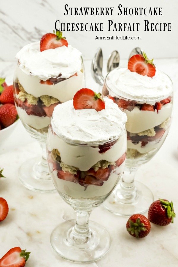 overhead view of three strawberry cheesecake partfait desserts on a white background, bowl of fresh strawberries to the left, individual strawberries on the white to the right, utensils in the  background