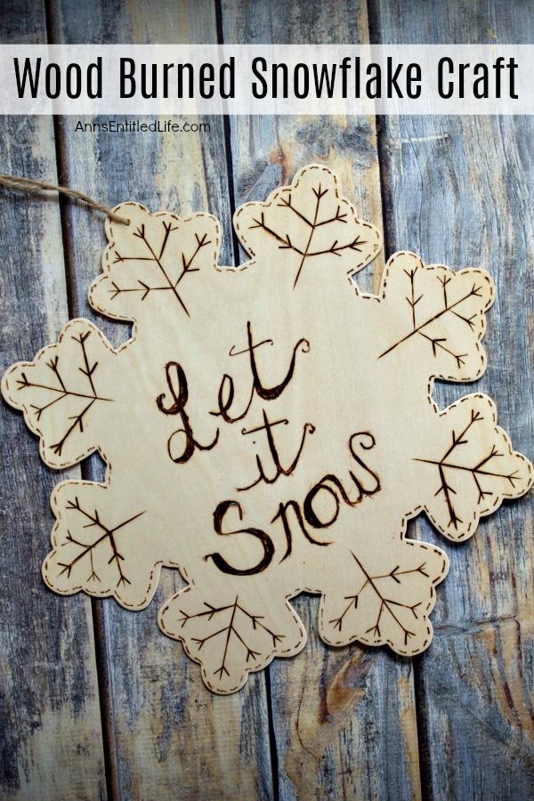 Wooden snowflake ornament etched with dashes, branches, and the words Let It Snow. There is a twine hanger at the end. This sits on top of a rustic blue surface.