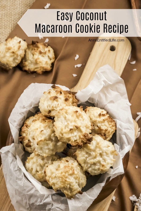 Overhead view of a wooden bowl of baked coconut macaroon cookies on a wooden server, all on a brown cloth. There are two more coconut macaroons on the cloth on the upper left side.
