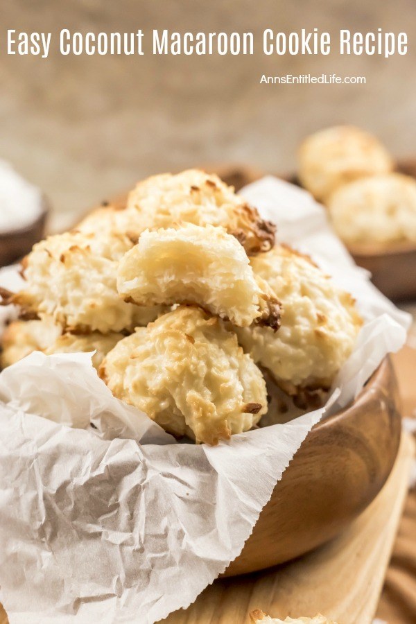 Wooden bowl of baked coconut macaroon cookies piled high, the top cookie has a bite of cookie removed. There are additional cookies in the blurred background.