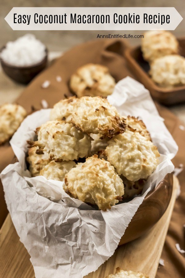 Wooden bowl of baked coconut macaroon cookies on a wooden server on top of a brown cloth. There are two additional cookies and a small bowl of shredded cocobut in the upper left