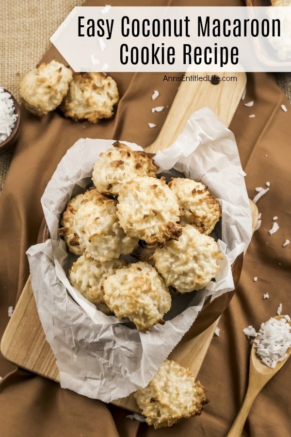 Overhead view of a wooden bowl of baked coconut macaroon cookies on a wooden server, coconut in a bowl in the upper left background, more coconut macaroons in a tray in the upper right background