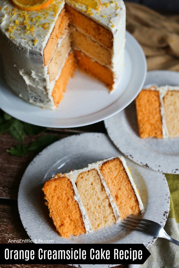 Two pieces of plated orange creamsicle cake, the remaining three-tier cake is on a white cake plate behind the pieces of served cake