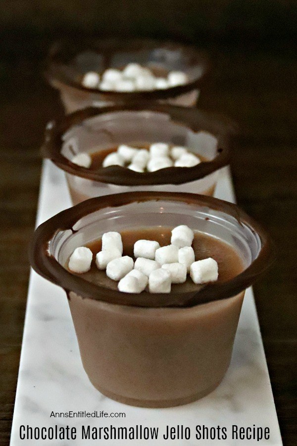 Three chocolate marshmallow jello shots in a line on top of a marble platter against a dark background.