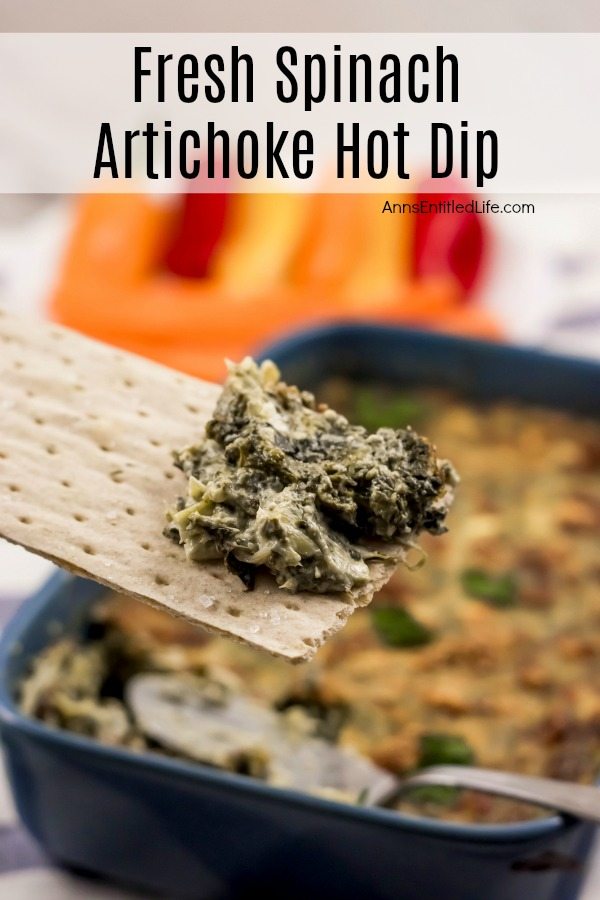 Close up of a serving of spinach artichoke dip on a cracker, a blue pan filled with the remaining baked spinach artichoke dip is in the background