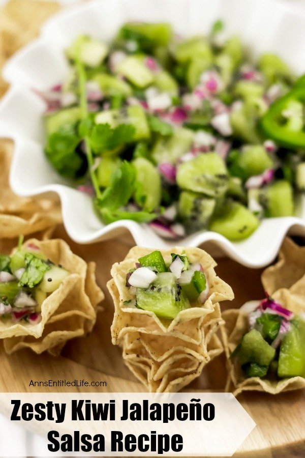  white bowl filled with kiwi fruit jalapeño salsa surrounded by tortilla chips on a cutting board. There are stacks of tortilla scoops in the front the top of each stack is filled with kiwi salsa