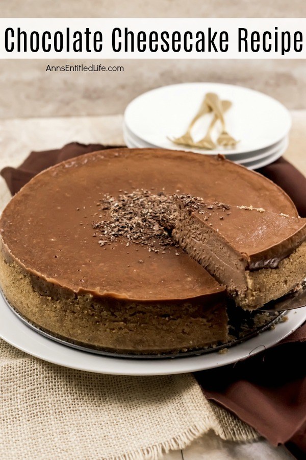 A whole chocolate cheesecake on a white serving plate. One piece is being lifted on a server, white cake plates with gold serving utensils are in the background.