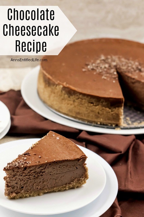 And piece of chocolate cheesecake on a white serving plate, the remainder of the chocolate cheesecake on a white serving plate in the background, all on top of a brown fabric.