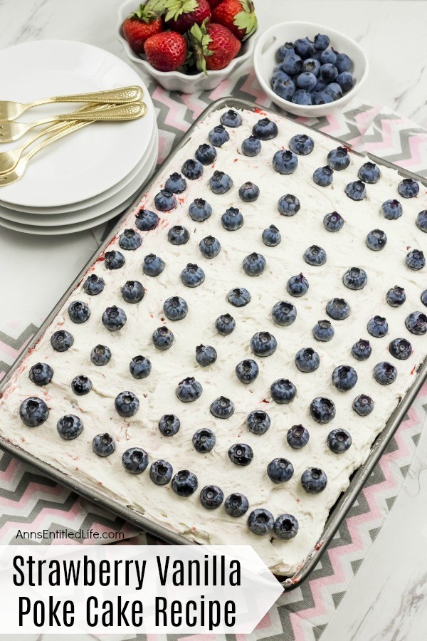 A 13x9 pan of strawberry vanilla poke cake decorated with fresh blueberries is in the center of the image. In the back, on the left, is a stack of white plates with gold silverware on top, in the middle back, a bowl of fresh strawberries, and the back right has a bowl of fresh blueberries