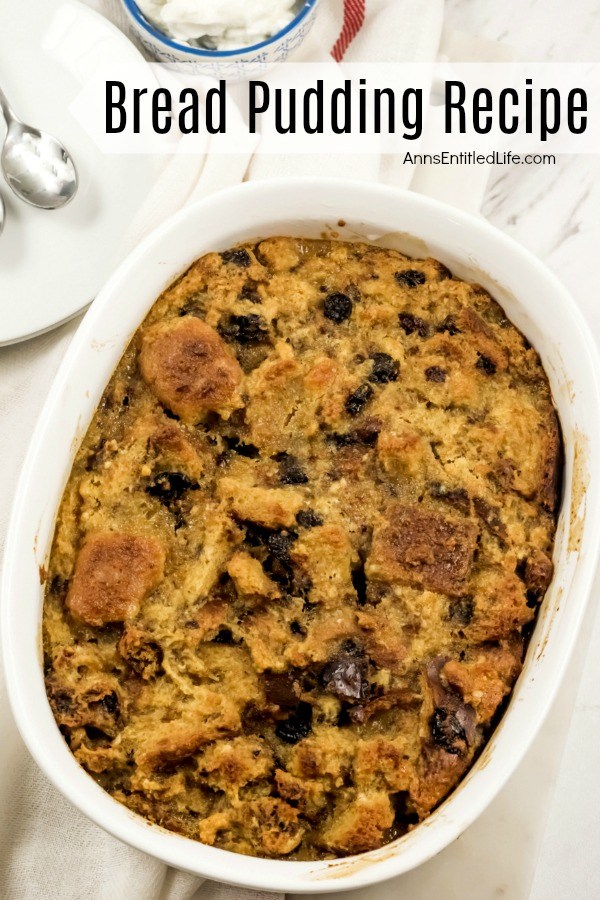 A white casserole bowl filled with baked bread pudding. There is a small bowl of whipped cream above the bread pudding, a white dish, and silverware to the left, all on top of a white napkin.