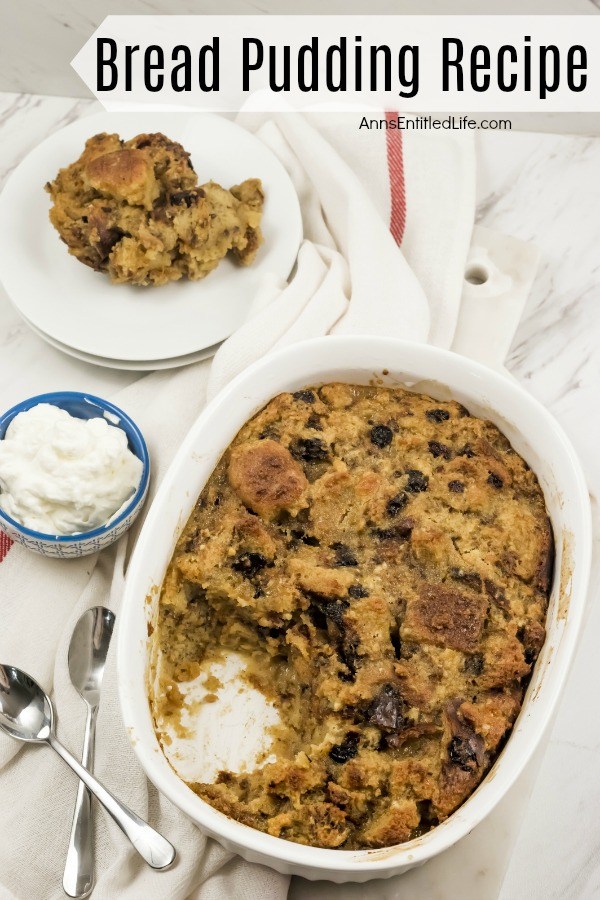 A white casserole bowl filled with baked bread pudding, one serving removed. There is a small bowl of whipped cream to the left bread pudding, above that sits a white dish filled with the bread pudding serving, all on top of a white napkin.