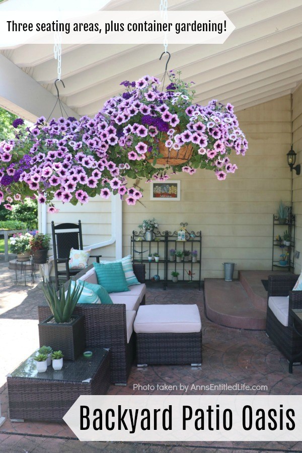 A backyard seating area with couch, and ottoman, chair, and rocking chair. There is an end table with a faux plant and three shelves with various floral. There are planters of flowers in the upper left, two purple petunia hanging baskets are in the foreground.