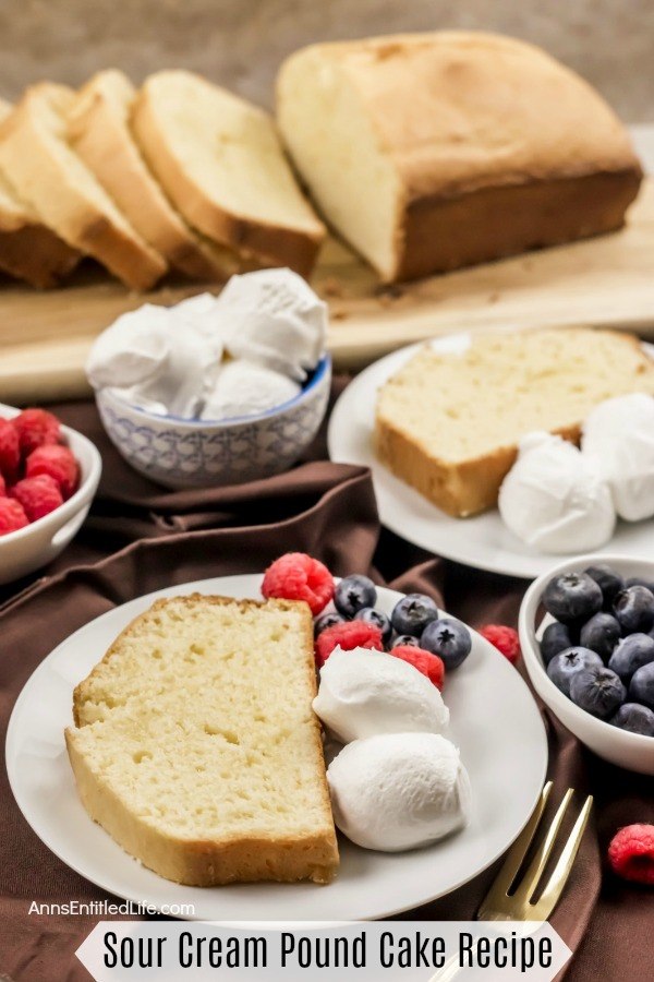 A white plate with a slice of sour cream pound cake, sour cream balls, blueberries and raspberries. A second such plate is in the upper right. Directly to the right is a bowl of blueberries. In the upper left are a bowl of raspberries and sour cream balls. At the top of the photo is the cut sour cream pound cake on a wooden cutting board.