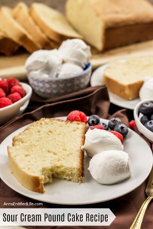 A white plate with a slice of sour cream pound cake with a bite removed, sour cream balls, blueberries and raspberries. A second such plate is in the upper right. Directly to the right is a bowl of blueberries. In the upper left are a bowl of raspberries and sour cream balls. At the top of the photo is the cut sour cream pound cake on a wooden cutting board.