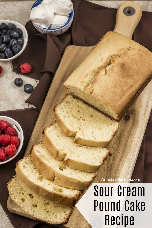 A sliced sour cream pound cake on a wooden cutting board. A bowl of raspberries to the left, a bowl of blueberry in the upper left, a bowl of source cream balls are above that, all on a dark-brown linen.