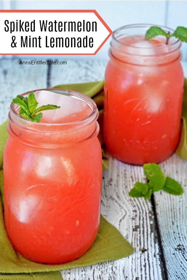 Two Mason jar glasses filled with watermelon and mint lemonade on top of a moss green napkin set on a distressed blue board. There is a spring of mint in from of one of the jars on the board.