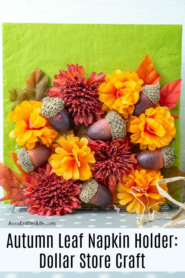 A plastic napkin holder decorated with faux leaves, flower heads, and acorns set against a blue background. There are green napkins in the holder.